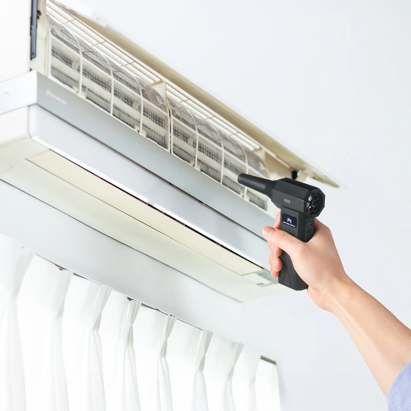 エアコンのフロントパネルが開かれ、埃がたまったフィルターが露出しています。手持ちの電動ブロワーを使って、フィルターの清掃を行っている様子を捉えた画像です。