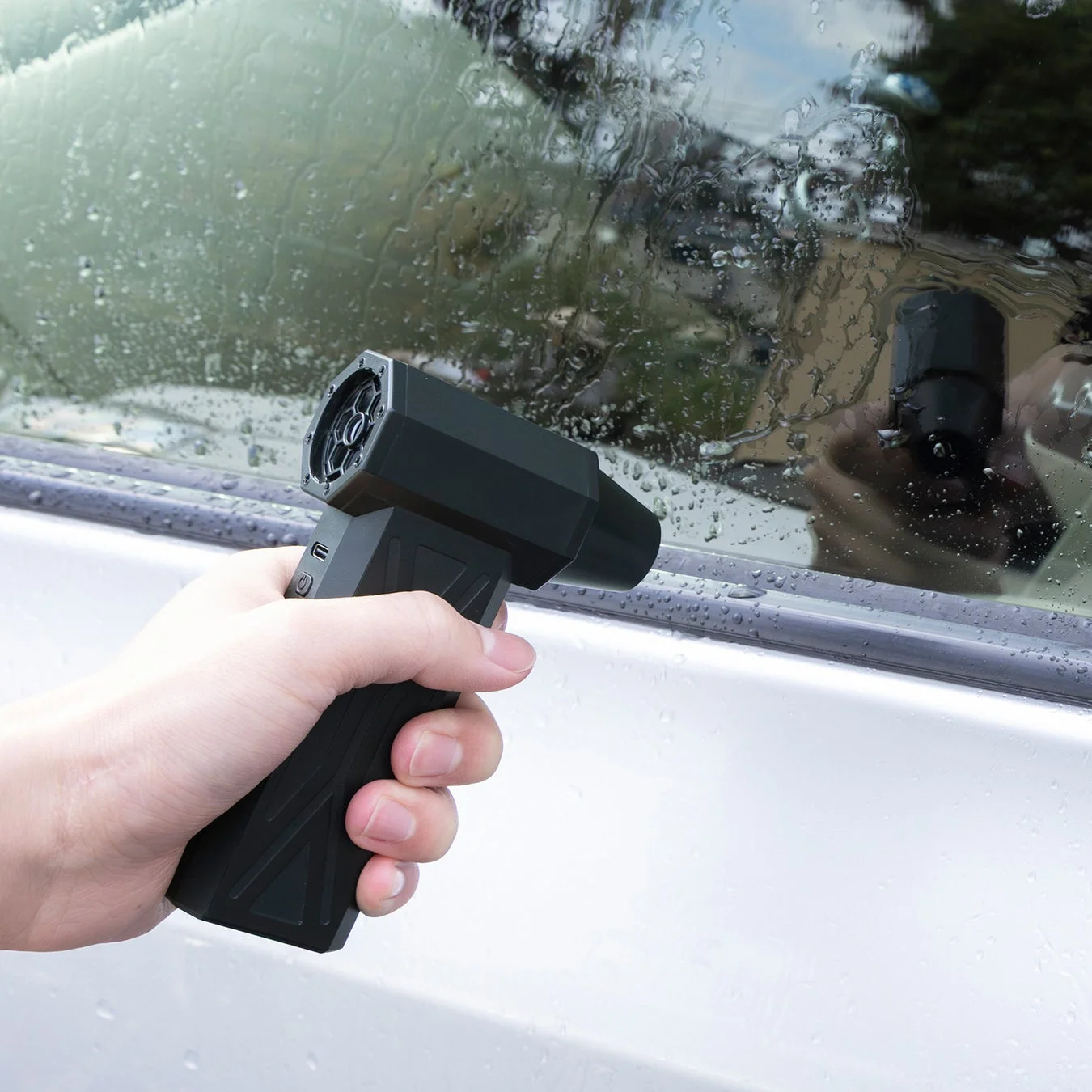手が黒いポータブルブロワーのようなデバイスで、水滴のついた車の窓を乾燥させている様子を捉えた画像です。